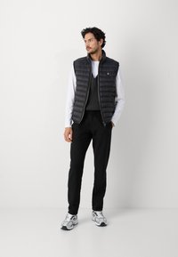 Zwarte puffer vest met ritssluiting, gedragen over een grijze gebreide trui, gecombineerd met zwarte broeken en zilveren sneakers. Minimalistisch ontwerp.