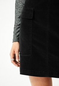 Musta corduroy-hame, jossa on etutasku ja ompelu yksityiskohtia. Tekstuuri on pehmeä ja sileä pinta. Käsissä on sormus.