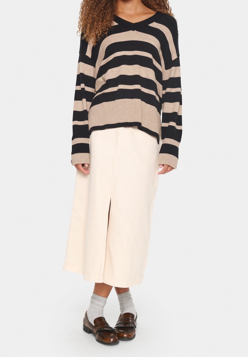 Femme portant un pull à rayures noires et beiges, une jupe midi crème avec une fente avant, des chaussettes grises et des mocassins en cuir marron, debout sur fond blanc.