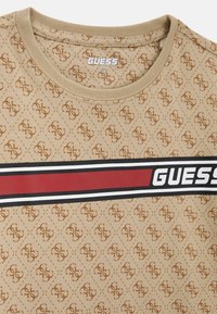 T-shirt beige avec un motif de logo répété, un col ras du cou, et une bande de marque noire et rouge audacieuse sur la poitrine. Tissu texturé.