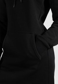 Gros plan d'une personne portant une robe sweat à capuche noire avec une poche kangourou devant et une main glissée dans la poche.