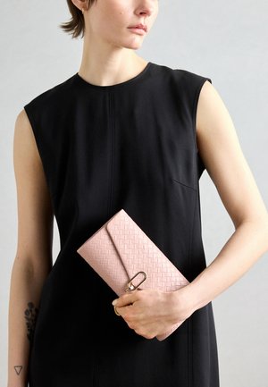 Nainen hihattomassa mustassa mekossa, joka pitää vaaleanpunaista kudottua kirjekuorilaukkua, jossa on kultainen lukko, yksinkertaisen harmaan taustan edessä.