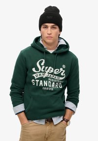 Sudadera verde con texto gráfico blanco, bolsillo canguro en la parte delantera, combinada con una camisa a rayas azules y blancas, y acompañada de un gorro de punto negro.