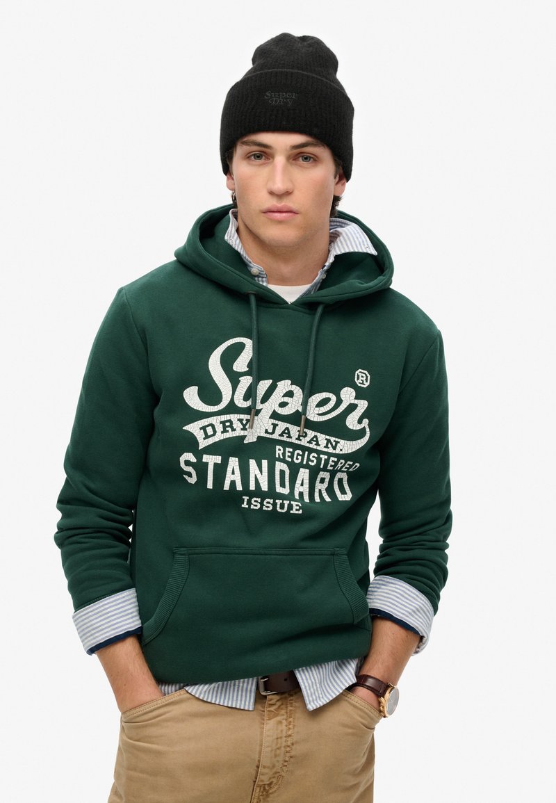 Sudadera verde con texto gráfico blanco, bolsillo canguro en la parte delantera, combinada con una camisa a rayas azules y blancas, y acompañada de un gorro de punto negro.