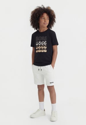 Copil cu păr creț purtând un tricou negru cu inscripția "BOSS", pantaloni scurți albi cu logo-ul "BOSS", șosete albe și adidași albi, stând cu mâinile în buzunare.