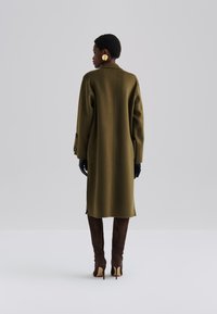 Abrigo de lana verde oliva con corte recto, aberturas laterales y puños abotonados. Combinado con botas de ante marrón hasta la rodilla y guantes negros.