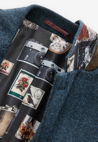 Blaues Wollblazer im Fischgrätenmuster mit einem kontrastierenden bedruckten Futter, das Kameras, Briefmarken und blumige Motive in gedämpften Farben zeigt.