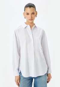 Camicia bianca a maniche lunghe con colletto a punta, taschino frontale, tessuto strutturato e leggera trasparenza, abbinata a jeans blu.