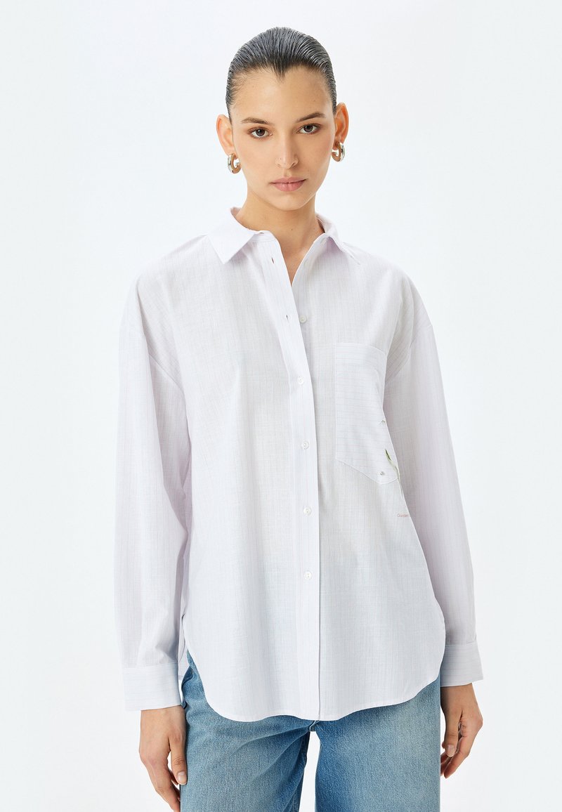 Camicia bianca a maniche lunghe con colletto a punta, taschino frontale, tessuto strutturato e leggera trasparenza, abbinata a jeans blu.