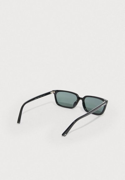 Svarta rektangulära solglasögon med gröna linser, slimmad plastbåge och smala svarta skalmar med subtil märkning på sidorna.