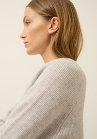 Hellgrauer Strickpullover mit weicher Textur, lockerer Passform und weitem Ausschnitt, an einer Person im Profil vor einem neutralen Hintergrund gezeigt.