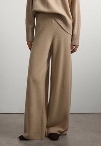 Pantalon en maille beige à jambes larges avec une texture douce, présentant une taille haute et un ourlet côtelé. Associé à des chaussures marron foncé.
