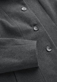 Ärmel und Vorderseite eines dunkelgrauen Wollmantels mit drei vertikal angeordneten schwarzen Knöpfen auf der Stoffoberfläche.