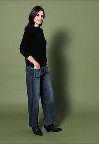 Czarny sweter, dżinsy niebieskie o wysokim stanie, szerokie nogawki, z przetarciami, z czarnymi botkami na obcasie sięgającymi kostki na zielonym tle.