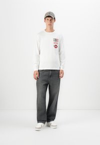 Young man wearing a gray cap, white long-sleeve shirt with red patches, dark gray wide-leg jeans, and white sneakers, standing against a white wall.