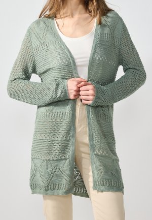 Frau trägt eine hellgrüne, offen gestrickte lange Strickjacke über einem weißen Oberteil und beigen Hosen und hält die Kanten der Strickjacke an der Taille geschlossen.