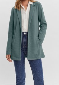 Femme portant un blazer bleu sarcelle sur une chemise blanche boutonnée et un jean bleu, debout avec une main dans la poche devant un fond uni.