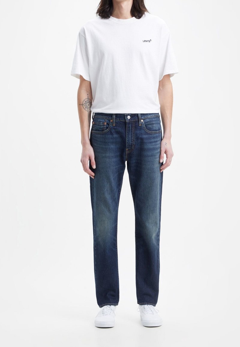 White cotton t-shirt with a small black Levi's logo, paired with dark blue denim jeans featuring a slight fade and straight-leg fit. White sneakers.