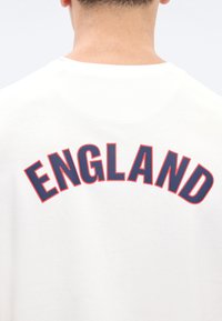 Άνθρωπος που φοράει λευκό πουκάμισο με τη λέξη «ENGLAND» εκτυπωμένη με μεγάλα σκούρα μπλε γράμματα με κόκκινο περίγραμμα στο πάνω μέρος της πλάτης.