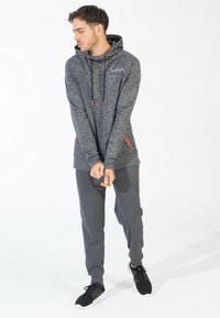 Felpa grigia con dettagli rossi e cordini, abbinata a pantaloni jogger grigi. L'outfit presenta un tessuto testurizzato e un design sportivo.