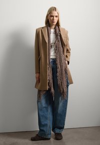 Manteau en laine beige au style ajusté, porté par-dessus une chemise blanche. Un pantalon en denim bleu ample et une écharpe en laine texturée marron. Chaussures sombres.
