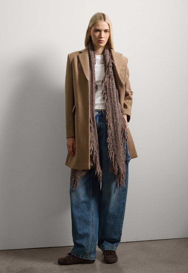 Manteau en laine beige au style ajusté, porté par-dessus une chemise blanche. Un pantalon en denim bleu ample et une écharpe en laine texturée marron. Chaussures sombres.
