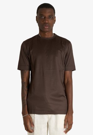 Giovane uomo con capelli corti indossa una t-shirt marrone a maniche corte e pantaloni bianchi, in piedi con le braccia rilassate ai lati.