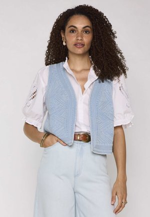 Femme aux cheveux bouclés portant un chemisier blanc aux manches brodées, un gilet texturé bleu clair, un pantalon taille haute et des accessoires dorés.