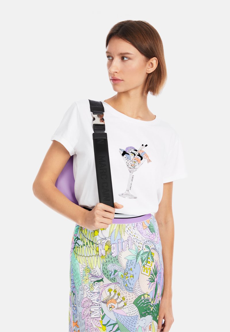 Weißes T-Shirt mit einer bunten Drink-Illustration auf der Brust, kombiniert mit einem gemusterten Rock in Pastellfarben und einer schwarzen Umhängetasche.