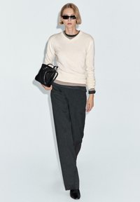 Kvinde med kort blondt hår iført sorte solbriller, cremefarvet sweater, mørkebukser, sorte støvler, bærende på sort håndtaske, gående mod en enkel baggrund.