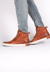 Zapatos de cuero marrón de caña alta con acabado texturizado, suelas de goma blanca y detalles de cordones grises, combinados con un denim azul claro.