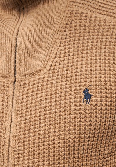 Μπεζ πλεκτό πουλόβερ με υφή, με κεντρικό φερμουάρ και ένα ναυτικό μπλε λογότυπο αλόγου στη αριστερή πλευρά.