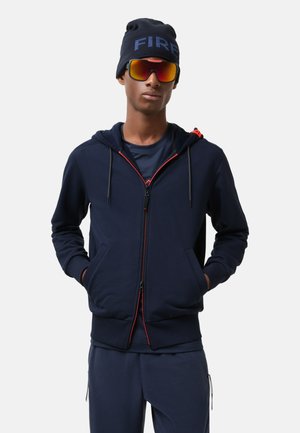 Navy zip-up hoodie with red accents, drawstring hood, two front pockets, paired with a matching outfit, beanie with "FIRE," and sunglasses.