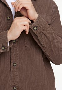 Camisa de pana marrón con cierre de botones en la parte delantera, que cuenta con un bolsillo en el pecho y una textura acanalada. Las mangas están arremangadas, mostrando los detalles de los botones.