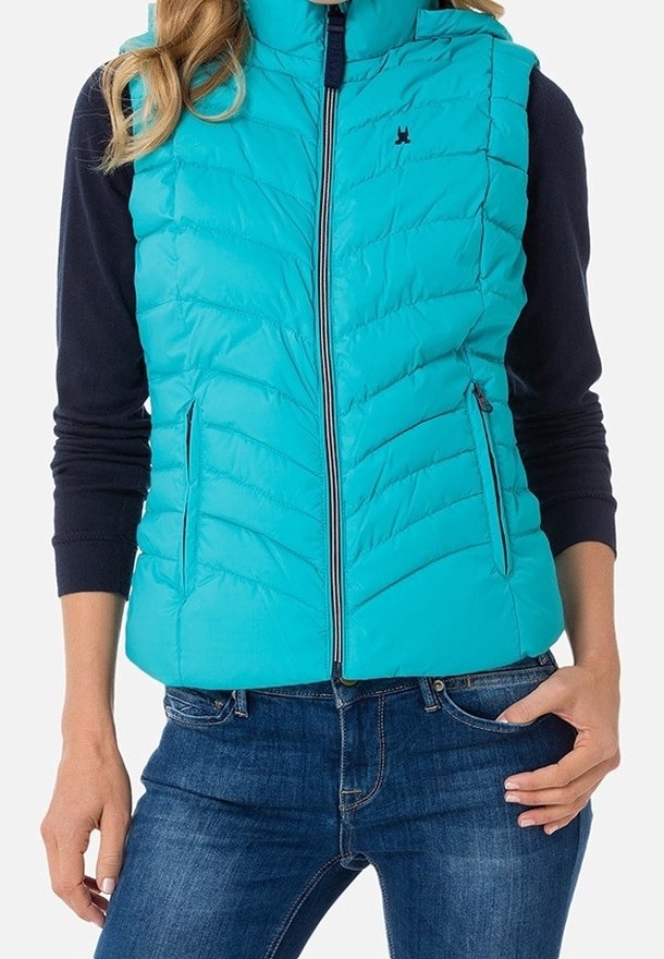 Femme portant une veste matelassée turquoise sans manches par-dessus un chemisier bleu marine à manches longues et un jean bleu, avec une main dans la poche.