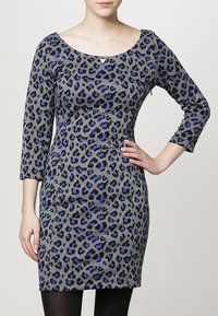 Robe à imprimé léopard gris avec des accents bleus, manches trois quarts et décolleté arrondi, fabriquée dans un tissu extensible. Détail subtil en forme de triangle.