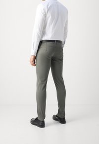 Pantaloni slim-fit verde oliva realizzati in cotone con una texture liscia. Presentano tasche posteriori e una cintura nera. Indossati con una camicia bianca con bottoni.