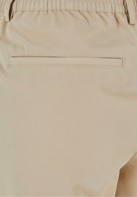 Detail tkaniny béžové barvy, který ukazuje zadní kapsu kalhot s jednou švovou kapsou a elastickým pasem.