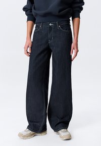 Jean en denim foncé à jambe large avec une taille haute, présentant des coutures visibles, une fermeture à bouton, ainsi que des poches avant et arrière.