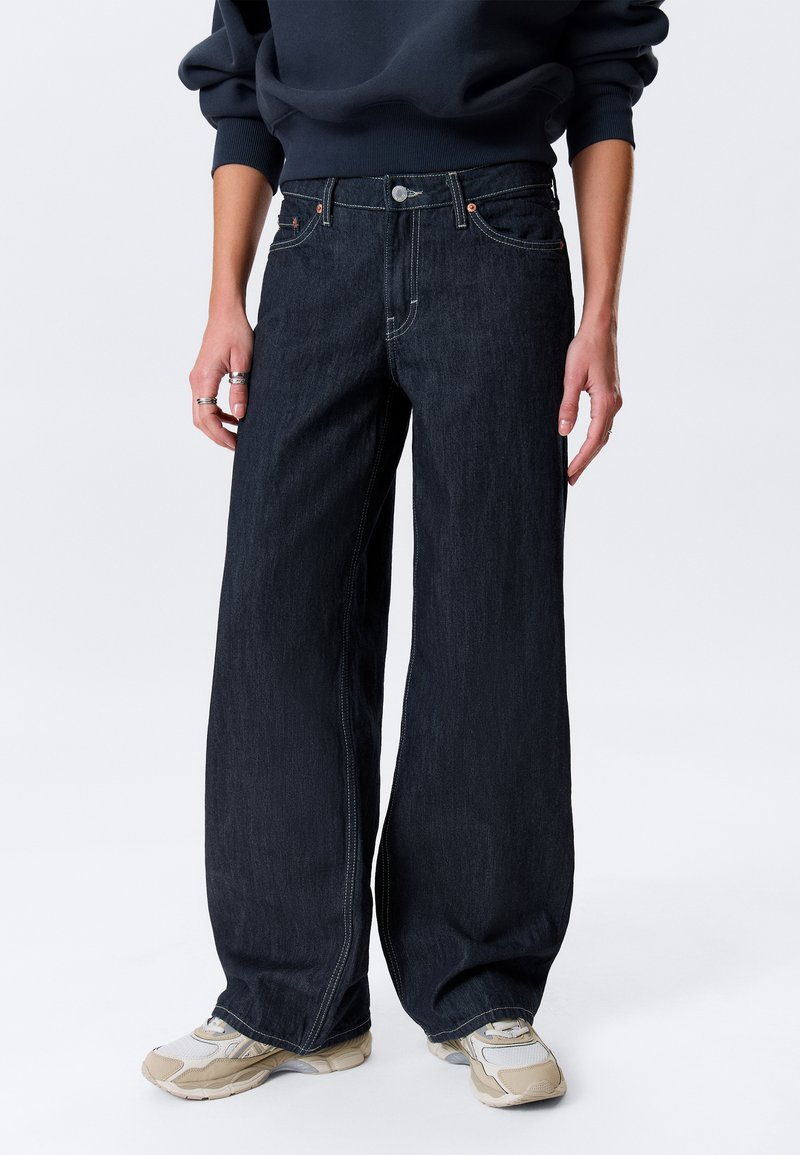 Jean en denim foncé à jambe large avec une taille haute, présentant des coutures visibles, une fermeture à bouton, ainsi que des poches avant et arrière.