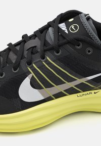 Fekete és szürke sportcipő, texturált felsőrésszel, sárga középtalppal és fehér Nike logóval. Fekete fűzőkkel és csíkos díszítésekkel rendelkezik.