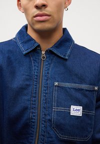 Donkerblauwe denim werkjack met een rits aan de voorkant, kraag en een borstzak met een witte "Lee" label. Contraststiksels.