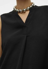 Top sans manches noir avec une finition texturée subtile, présentant un décolleté en V et accompagné d'un collier de perles métalliques imposant.