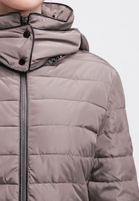 Puchowa kurtka w miękkim taupe z wysokim kołnierzem, błyszczącym czarnym zamkiem i zatrzaskami. Posiada poziome przeszycia dla uzyskania tekstury.