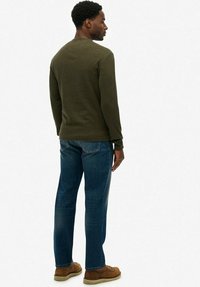 Oliwkowa koszula z długim rękawem o dzianinowej fakturze, w połączeniu z niebieskimi dżinsami. Brązowe buty sznurowane i widoczne przeszycia na spodniach.