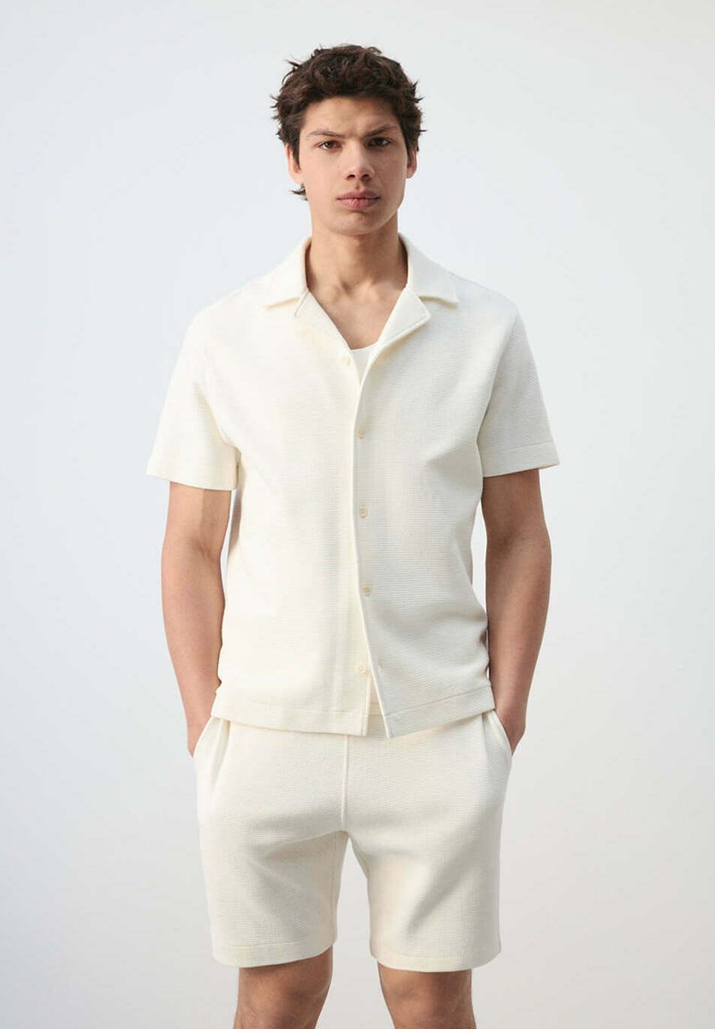 Joven de pie con las manos en los bolsillos, vistiendo una camisa de manga corta color blanco roto con botones y pantalones cortos a juego sobre un fondo liso.