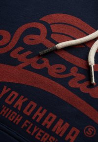 Donkerblauwe hoodie met een groot rood grafisch ontwerp, een gestructureerde stof en crèmekleurige trekkoorden met metalen uiteinden.