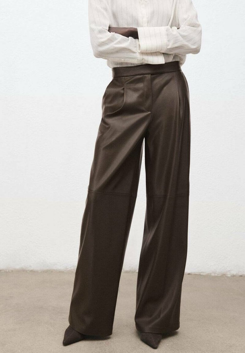 Femme debout les bras croisés, portant un pantalon en cuir marron foncé à taille haute et jambes larges, ainsi qu'une chemise beige à manches longues rentrée.