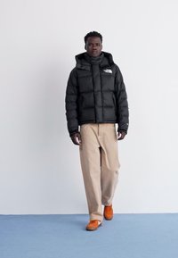 Svart puffjacka med hög krage och logotyp, kombinerad med beige vida byxor och orange sneakers på ett blått golv.