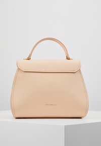 Beige läderhandväska med topphandtag, lockstängning, guldfärgad hårdvara och "COCCINELLE"-logotyp centrerad på framsidan, visad på en vit pelare.